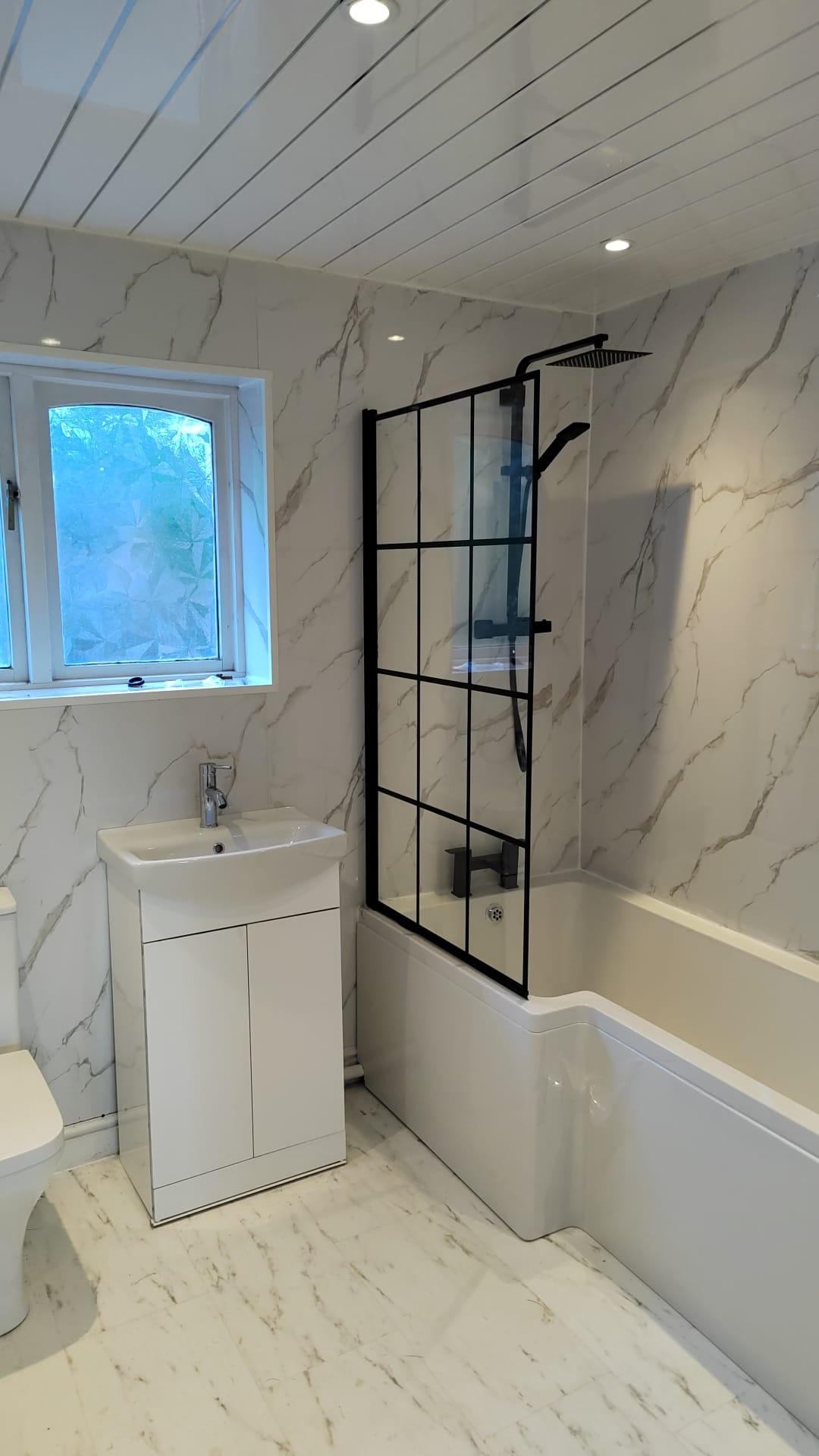 Refitted bathroom with marble-effect wall panels, black framed shower screen, black taps and a white vanity unit